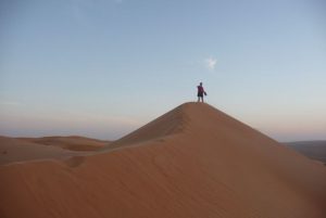 A man stands at the top of an orange sand dune as the sun begins to set in the background