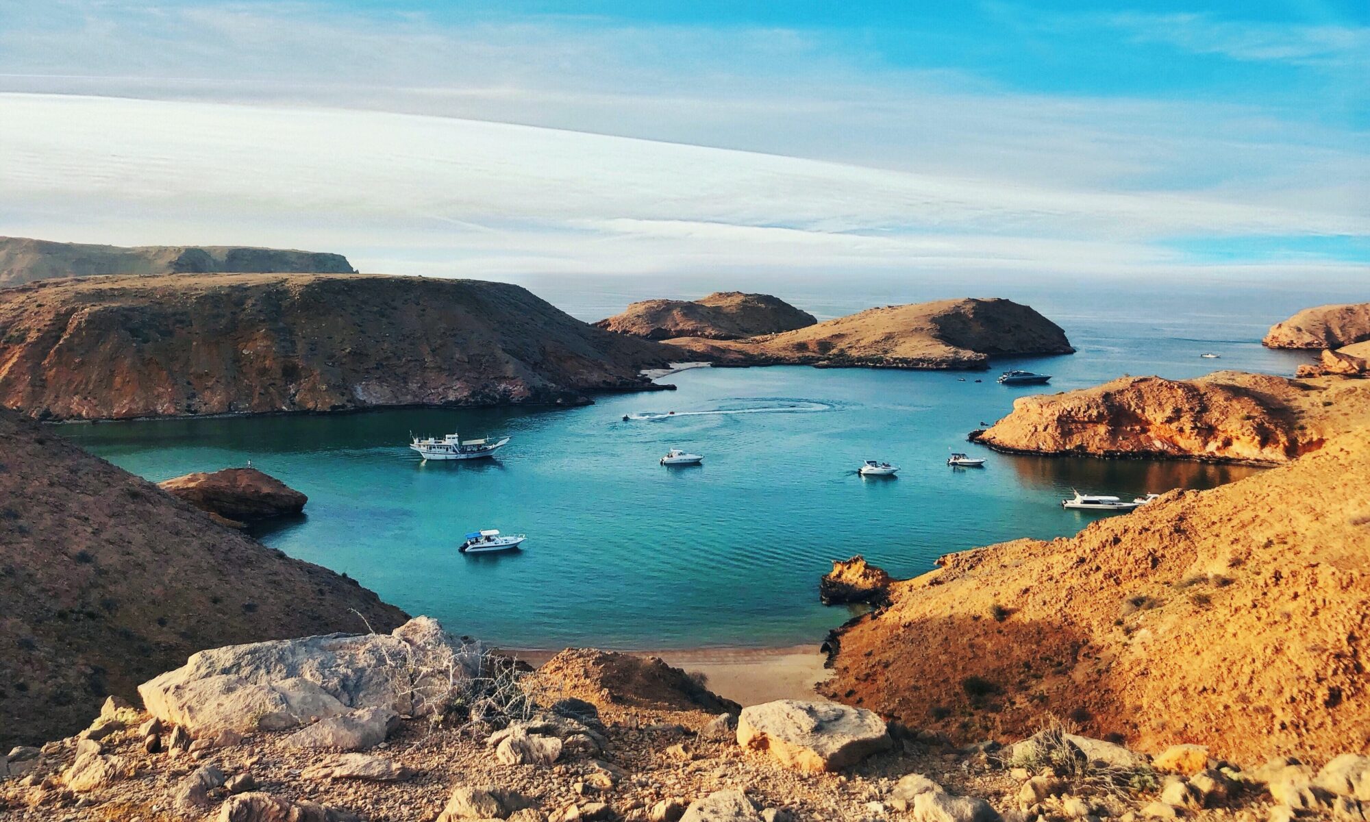 a brown and green rock formation on blue sea under blue sky during daytime, ships in the natural harbour, Oman