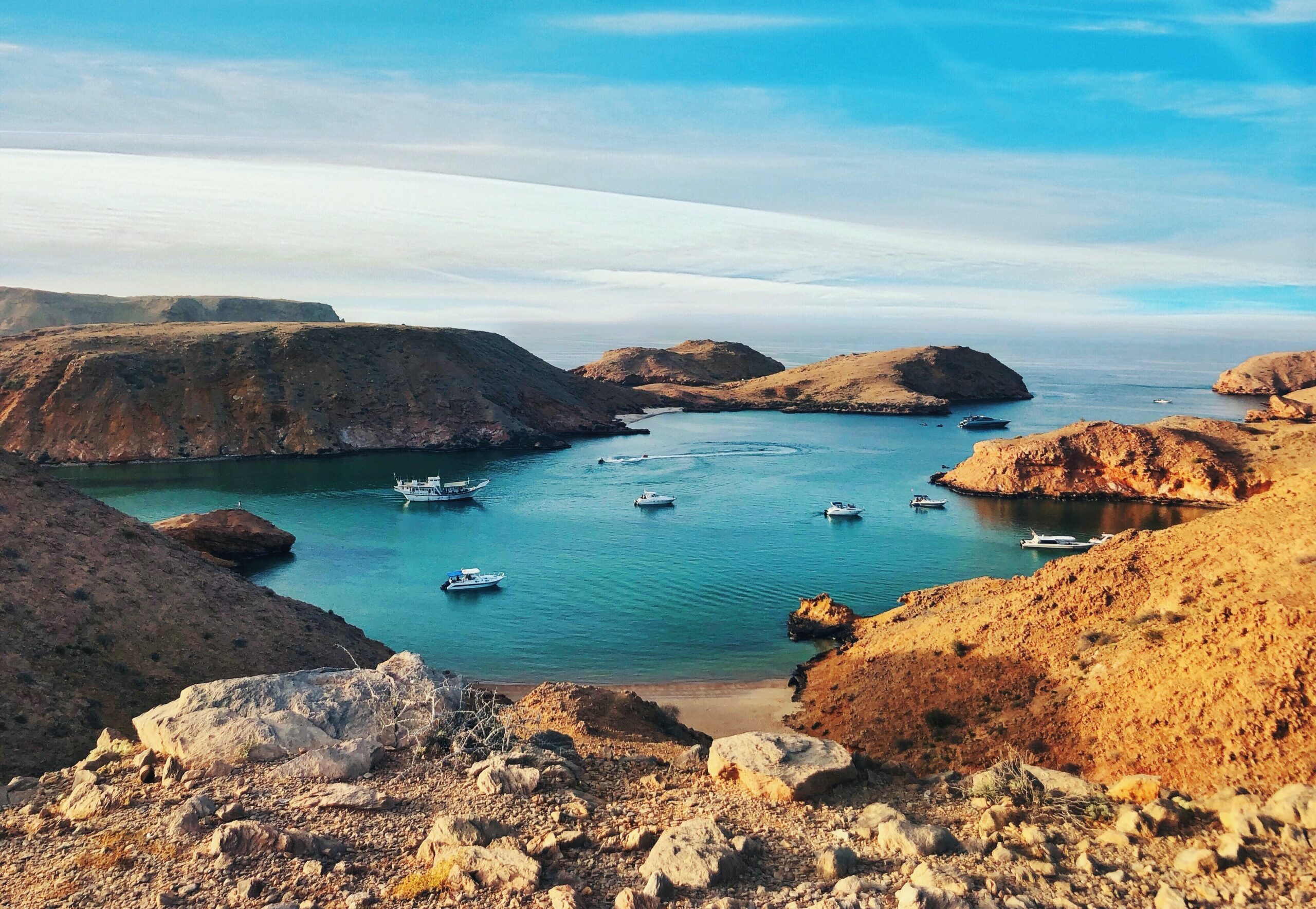 a brown and green rock formation on blue sea under blue sky during daytime, ships in the natural harbour, Oman